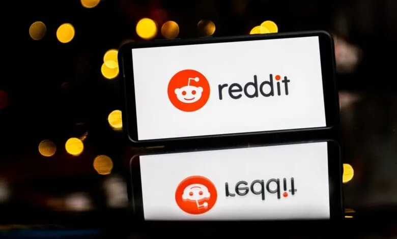 Smartphone displaying the Reddit logo: an orange alien mascot and 'reddit' in black text. Reflected on a dark surface.