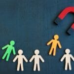 A red horseshoe magnet pulls an orange figure from a group of beige and green figures on a dark blue background.