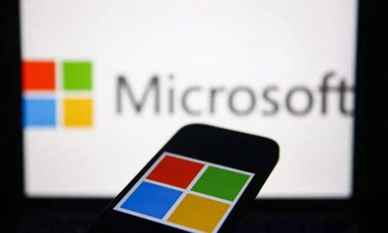 Close-up of a smartphone displaying the Microsoft logo, with a blurred computer screen in the background showing the same logo.