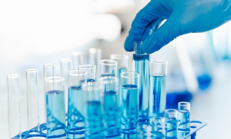A gloved hand places a test tube filled with light blue liquid into a rack of similar tubes.