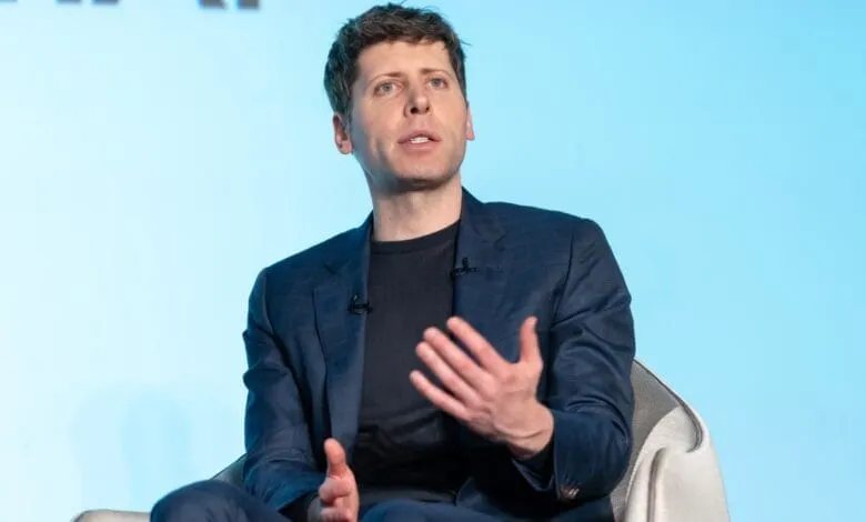 Sam Altman, CEO of OpenAI, in a dark blue blazer gestures while speaking at a conference. Light blue background.