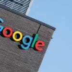The colorful Google logo is prominently displayed on the dark brick exterior of a building against a clear blue sky.