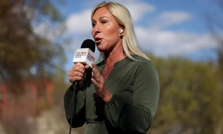A blonde woman in a green dress speaks into a Real America's Voice microphone outdoors on a sunny day.