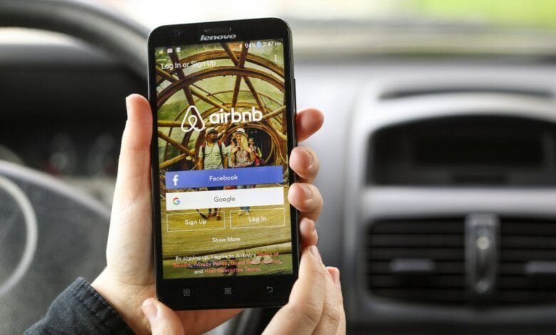 Hands hold a smartphone displaying the Airbnb login screen, offering Facebook and Google login options. A blurred car interior is in the background.