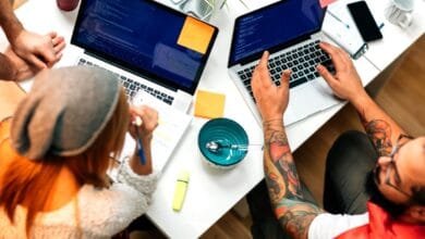 Overhead view of two people collaborating on laptops, code visible on screens. One person has tattoos.
