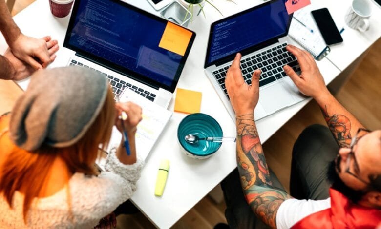 Overhead view of two people collaborating on laptops, code visible on screens. One person has tattoos.