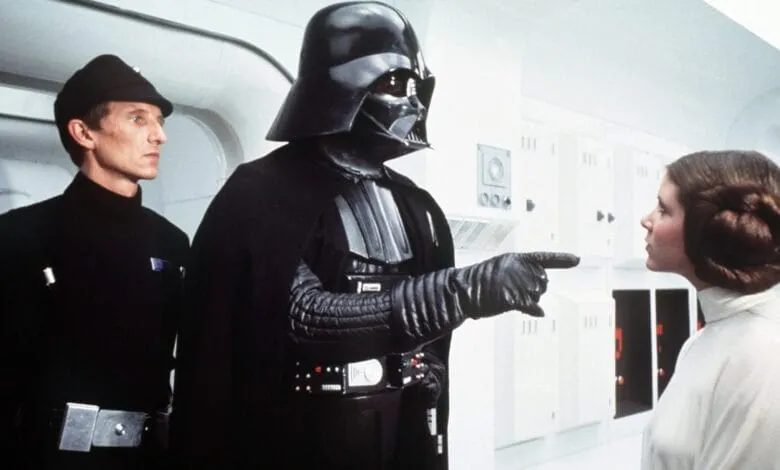 Darth Vader points at Princess Leia while an Imperial officer looks on in a stark white room.