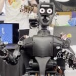 A black humanoid robot with articulated arms and hands interacts with a person's hand holding a device. A laptop displays a 3D animation.
