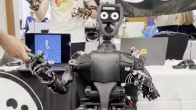 A black humanoid robot with articulated arms and hands interacts with a person's hand holding a device. A laptop displays a 3D animation.