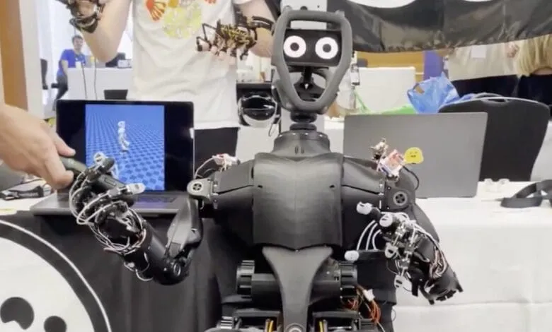 A black humanoid robot with articulated arms and hands interacts with a person's hand holding a device. A laptop displays a 3D animation.