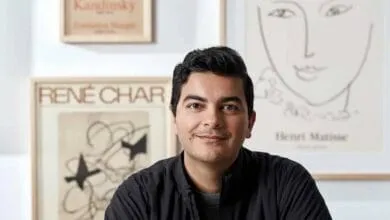 A man with dark hair and a dark jacket smiles gently. He's positioned in front of framed artwork with minimalist line drawings.