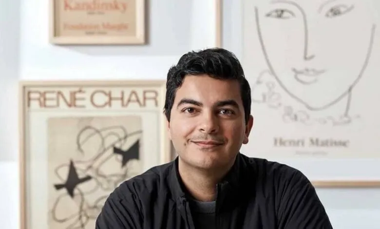 A man with dark hair and a dark jacket smiles gently. He's positioned in front of framed artwork with minimalist line drawings.
