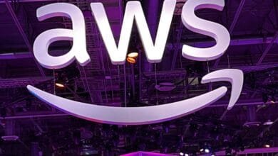 Large white AWS logo, including the Amazon smile, hangs from the ceiling of a purple-lit venue.