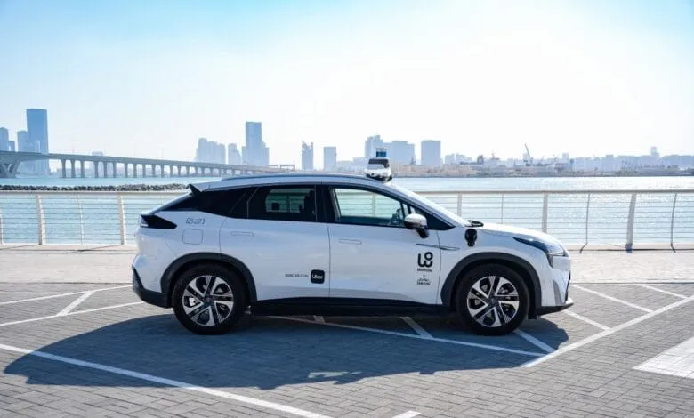 A white self-driving Uber WeRide vehicle parked by the water, with a city skyline in the background.