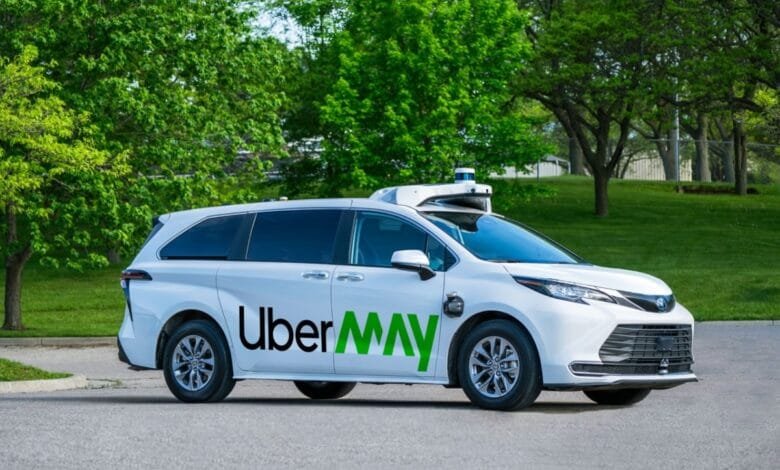 A white Uber MV self-driving minivan is parked in a lot with green trees in the background.