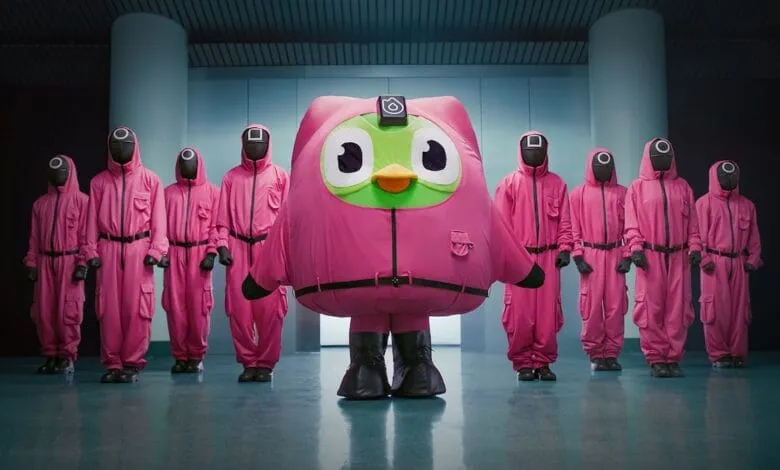 Duolingo's owl mascot in a pink Squid Game jumpsuit, flanked by figures in matching pink jumpsuits and black circle masks.