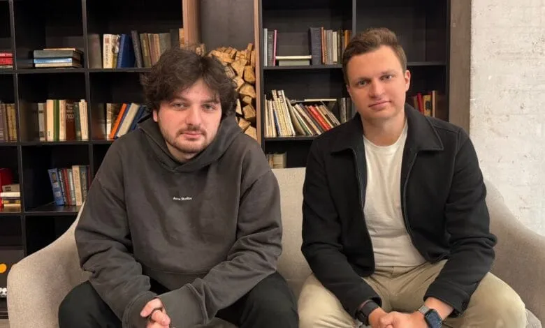 Two men, Rork founders, sit on a gray couch in front of a bookshelf, one wearing a dark hoodie and the other a black jacket.