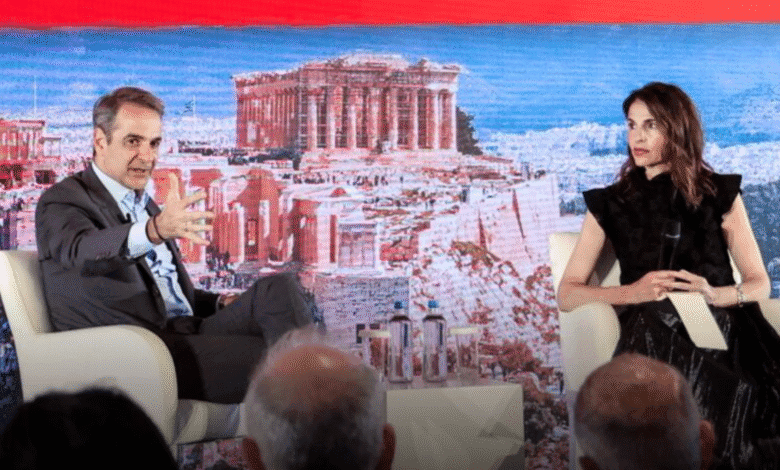 Greek Prime Minister Kyriakos Mitsotakis and a woman discuss on stage, with the Acropolis projected on a screen behind them.
