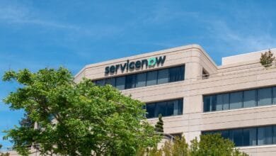 ServiceNow logo on a tan office building, lush green trees in the foreground, bright blue sky.