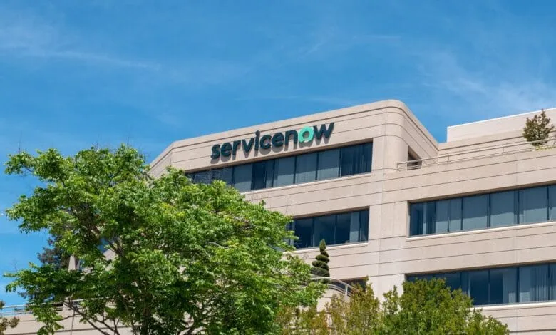 ServiceNow logo on a tan office building, lush green trees in the foreground, bright blue sky.