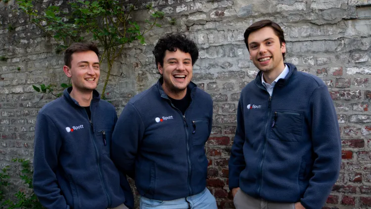 Three smiling men (Spott founders) in navy blue jackets stand against a brick wall. The jackets have a small logo.