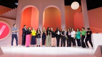 A large group of people, mostly women, stand on a stage, clapping and smiling. The backdrop is modern and minimalist.
