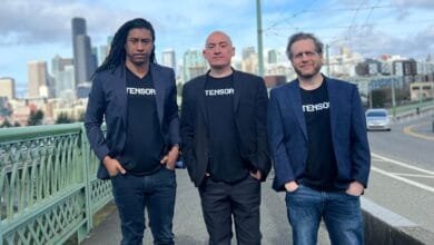 Three men in blazers and Tensor t-shirts stand on a bridge with a city skyline in the background.