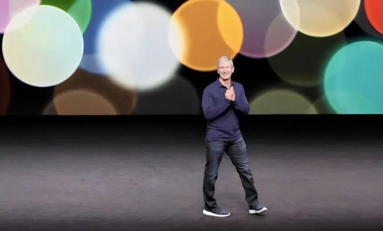 Tim Cook, Apple CEO, smiles on stage against a backdrop of colorful bokeh lights.