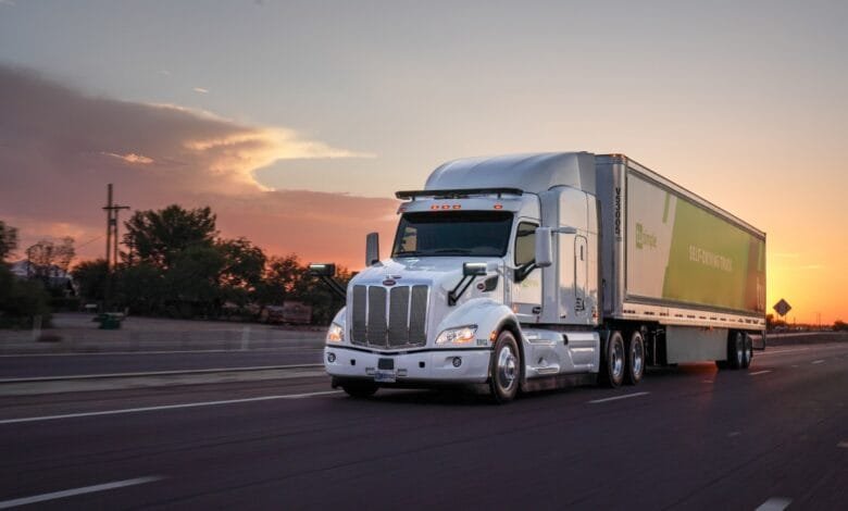 A white TuSimple self-driving truck speeds down a highway at sunset. The truck's trailer displays the company logo and 'Self-Driving Truck'.