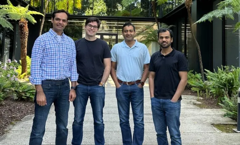 Four smiling men in casual attire stand outside a modern building. Lush greenery surrounds them.