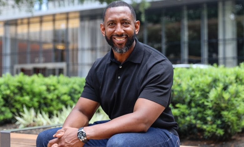 Smiling Black man sits on a park bench, wearing a black polo shirt and jeans.