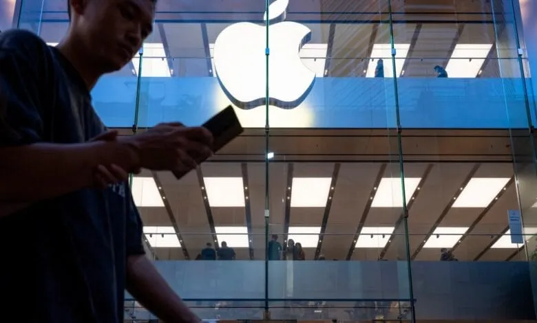 Silhouetted person uses phone outside brightly lit Apple store at dusk; iconic Apple logo glows.