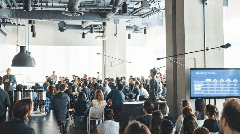 Large group of people attending a presentation in a modern office space. A screen displays data.
