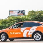 An orange Tesla Model X, branded with the GoCarma logo, drives past a billboard advertising GoCarma's HOV discounts.