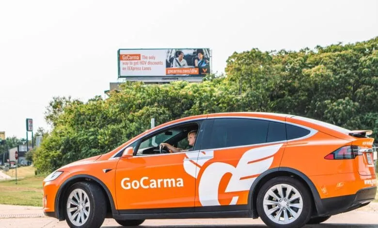 An orange Tesla Model X, branded with the GoCarma logo, drives past a billboard advertising GoCarma's HOV discounts.