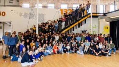 Large diverse group of smiling people pose for a photo on and around a staircase. A large wall graphic with numbers and a mountain design is behind them.