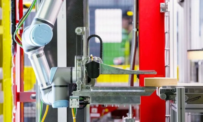 A robotic arm in a factory setting carefully places a cardboard box onto a conveyor belt. The arm is light blue and silver.