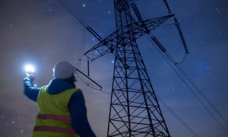 A lone worker silhouetted against a starry night sky, uses a flashlight while inspecting a large electrical pylon.