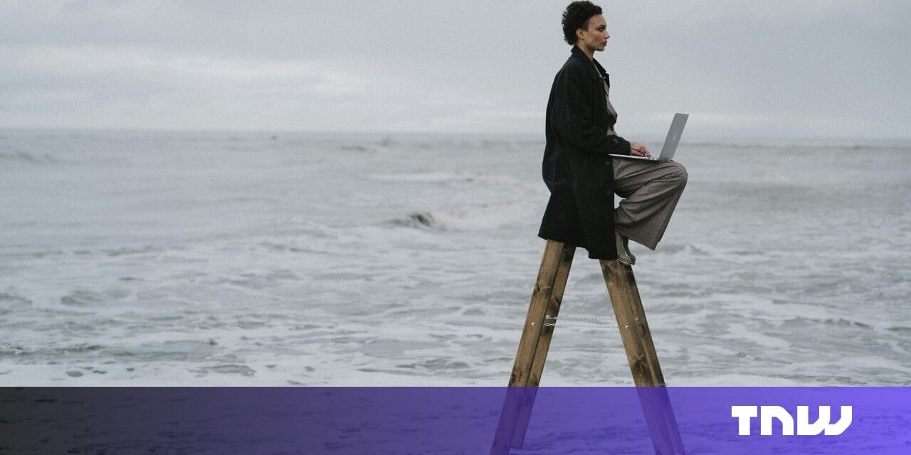 Woman sits on a wooden ladder by the ocean, working on a laptop. A moody, contemplative scene.