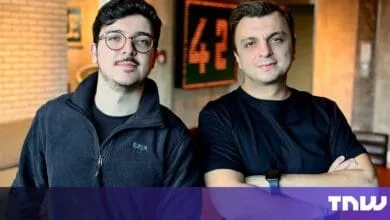 Two men, one wearing glasses, stand against a brick wall. They're dressed in dark clothing and have serious expressions.