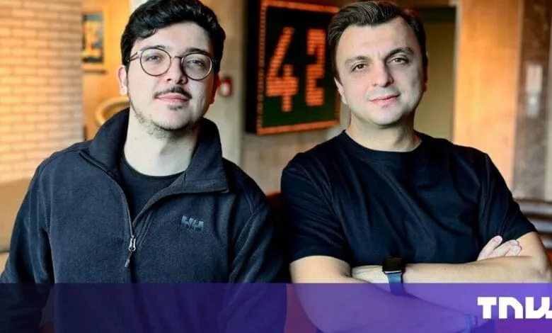 Two men, one wearing glasses, stand against a brick wall. They're dressed in dark clothing and have serious expressions.