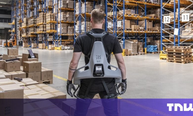 Warehouse worker wearing a grey exoskeleton, standing amidst stacked pallets and boxes.