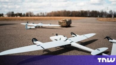 Three white drones on a tarmac, one in focus, others blurred in the background. A camouflaged object is visible further back.