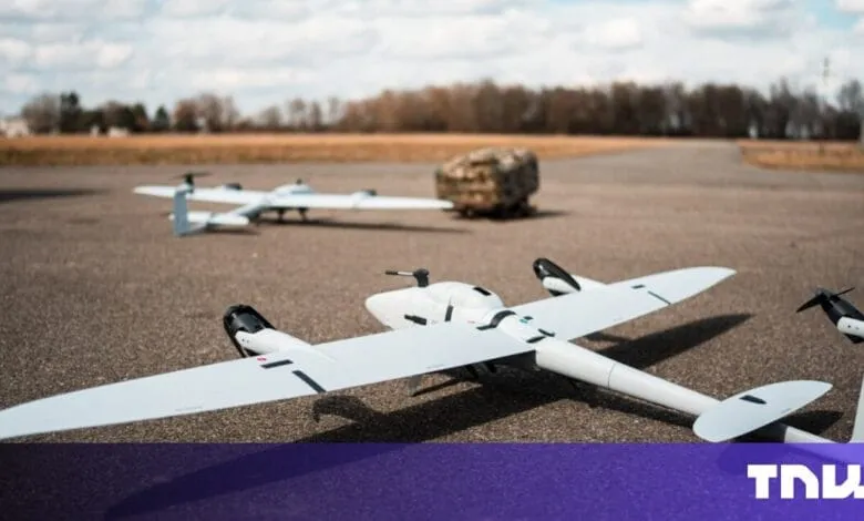 Three white drones on a tarmac, one in focus, others blurred in the background. A camouflaged object is visible further back.