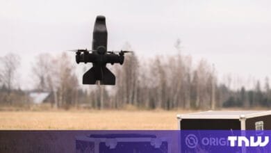 Black drone in flight over a field, approaching a dark-colored transport case.