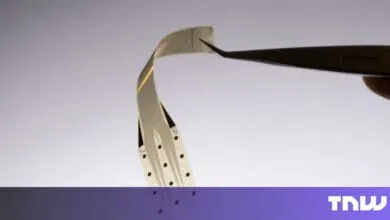 Close-up of a flexible, thin electronic circuit held by tweezers against a light gray background.