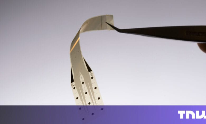 Close-up of a flexible, thin electronic circuit held by tweezers against a light gray background.