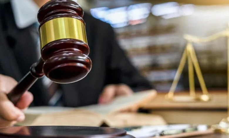 Close-up of a judge's gavel held in hand, with a blurred background of legal documents and scales of justice.