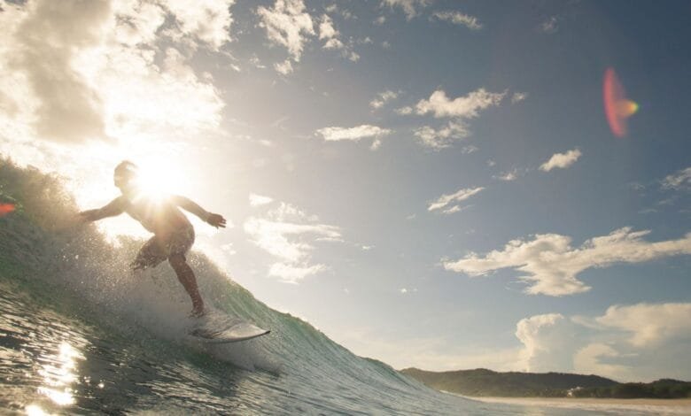 Silhouetted surfer rides a wave, sun shining behind them, creating a bright halo. Ocean and sky are visible.