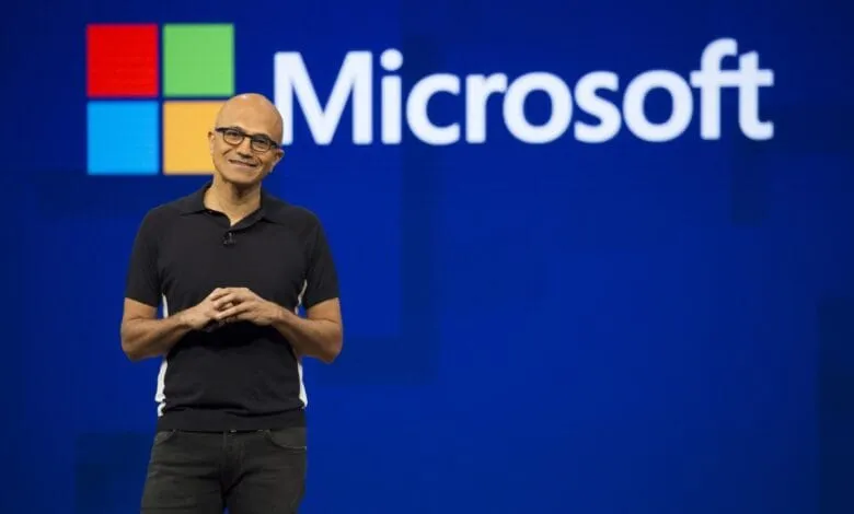 Satya Nadella, Microsoft CEO, smiles confidently on stage in front of a large Microsoft logo.